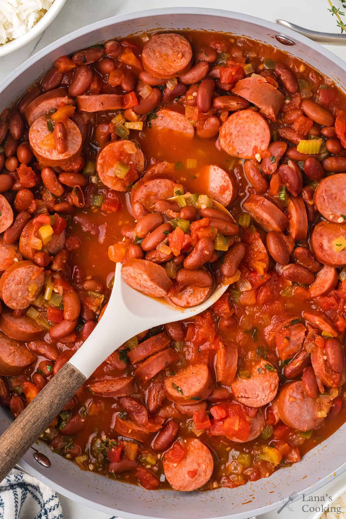 Sliced sausage, red beans, and vegetables in tomato sauce, with a spoon in a large white skillet.