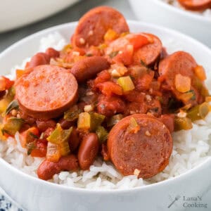 A bowl of white rice topped with sliced sausage, kidney beans, and a tomato-based vegetable sauce.