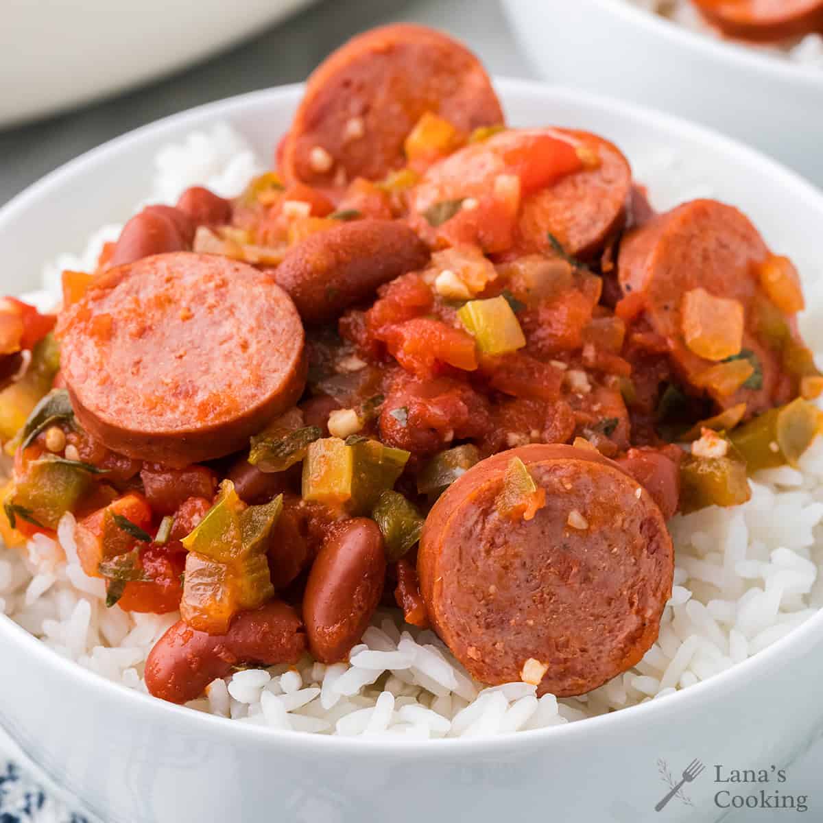 A bowl of white rice topped with sliced sausage, kidney beans, and a tomato-based vegetable sauce.