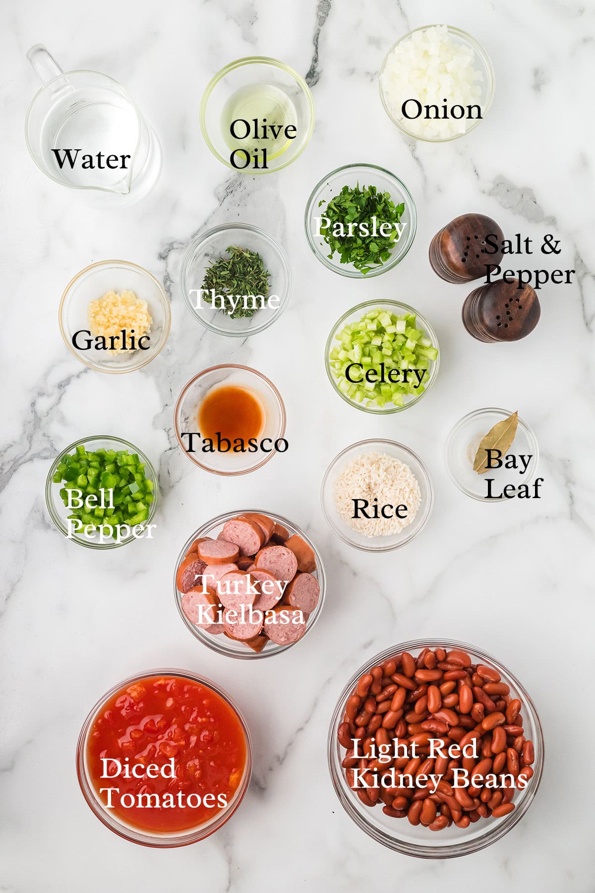 Bowls of labeled ingredients for red beans and rice, including veggies, spices, kielbasa, and beans on a white surface.