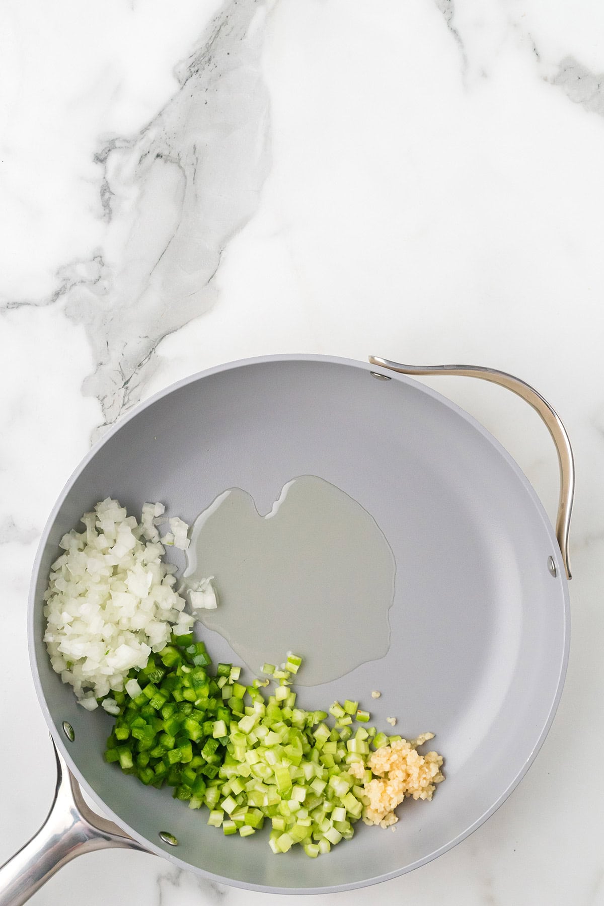 Gray skillet with oil, chopped onions, celery, green bell pepper, and minced garlic on a marble surface.