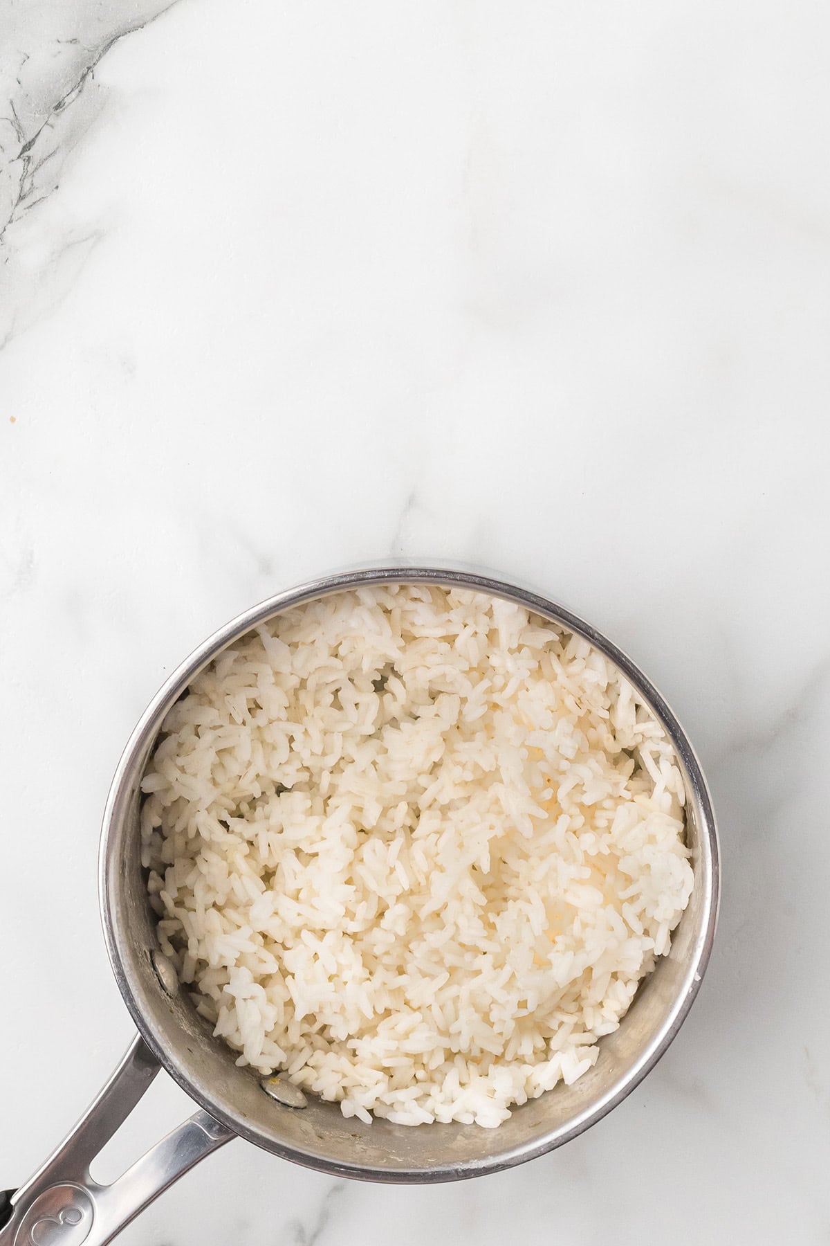 A saucepan filled with cooked white rice sits on a white marble surface.