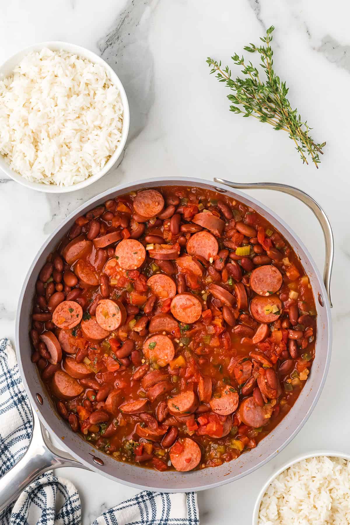A skillet of red beans and sausage in tomato sauce, with a bowl of white rice and fresh thyme beside it.