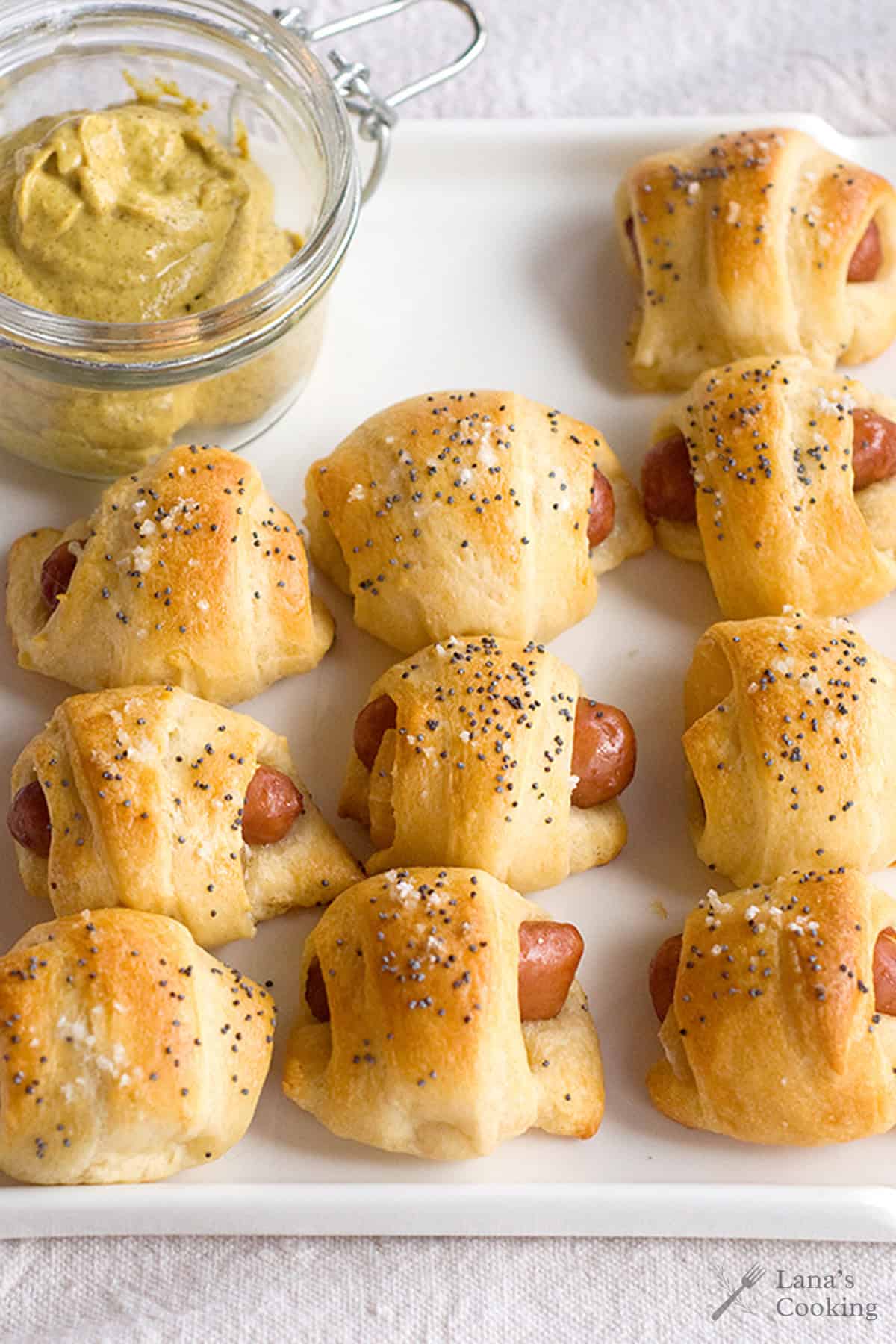 Mini pigs in blankets on a white plate with a jar of mustard dip in the corner.