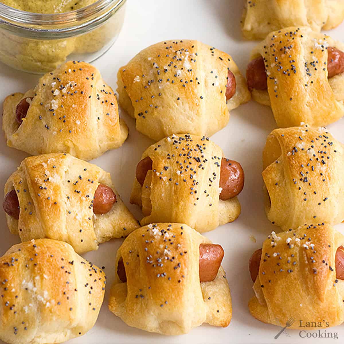 Mini pigs in a blanket topped with poppy seeds and salt on a white plate, with a jar of mustard in the corner.