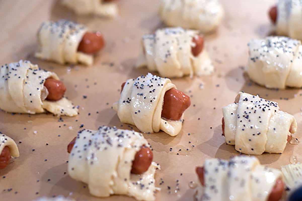 Mini sausages wrapped in dough, topped with poppy seeds, arranged on a baking sheet before baking.