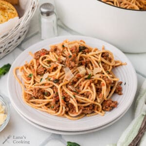 A plate of spaghetti with Bolognese sauce topped with grated cheese, set on a table with a salt shaker, a basket of bread, and a pot in the background.