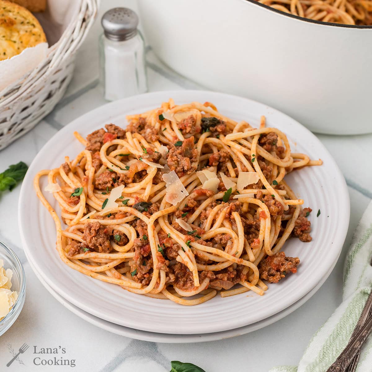 A plate of spaghetti with Bolognese sauce topped with grated cheese, set on a table with a salt shaker, a basket of bread, and a pot in the background.