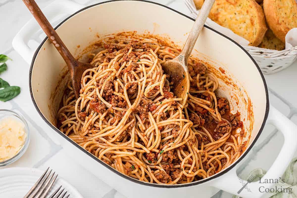 A pot of cooked spaghetti with Bolognese sauce is mixed with two wooden utensils, surrounded by forks, shredded cheese, basil leaves, and a basket of bread.