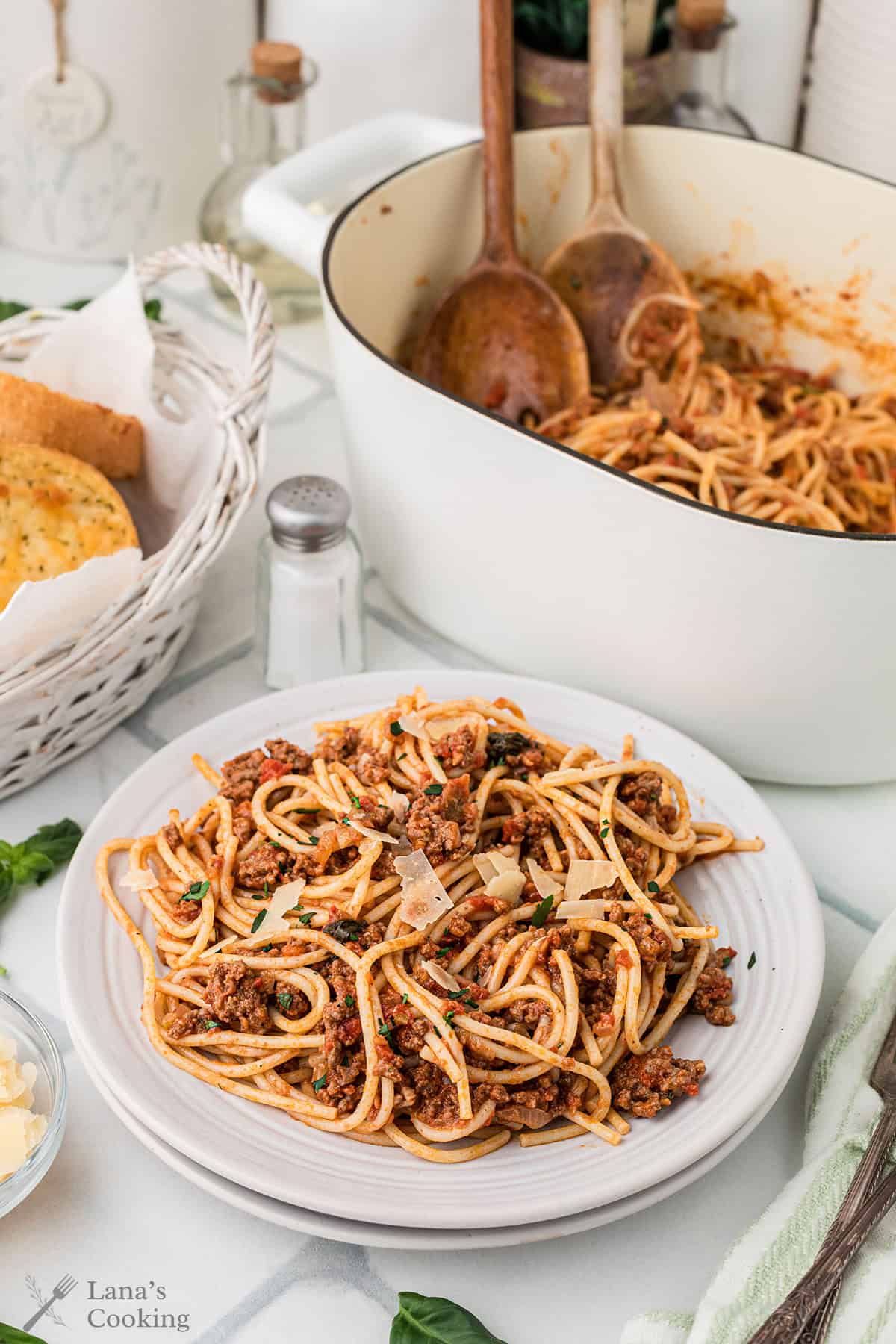 A plate of spaghetti with Bolognese sauce and parmesan sits next to a pot of pasta, wooden utensils, a bread basket, and a salt shaker on a white table.