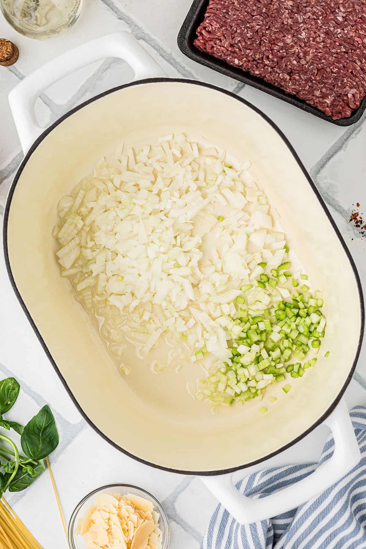 A white pot contains chopped onions and celery being saut&eacute;ed; nearby are raw ground meat, basil, parmesan, and other cooking ingredients.