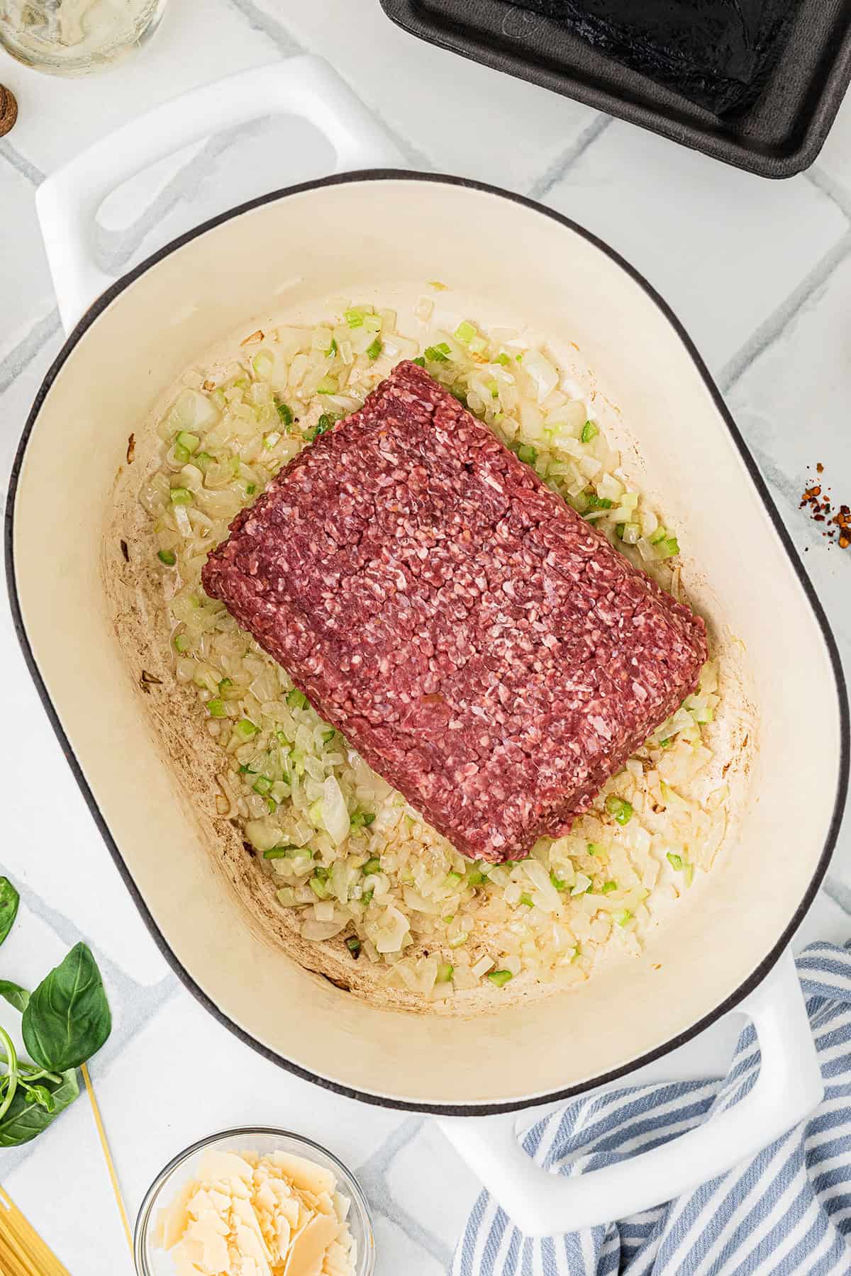 Ground beef sits atop saut&eacute;ed onions and celery in a white Dutch oven, surrounded by various cooking ingredients and utensils on a kitchen counter.