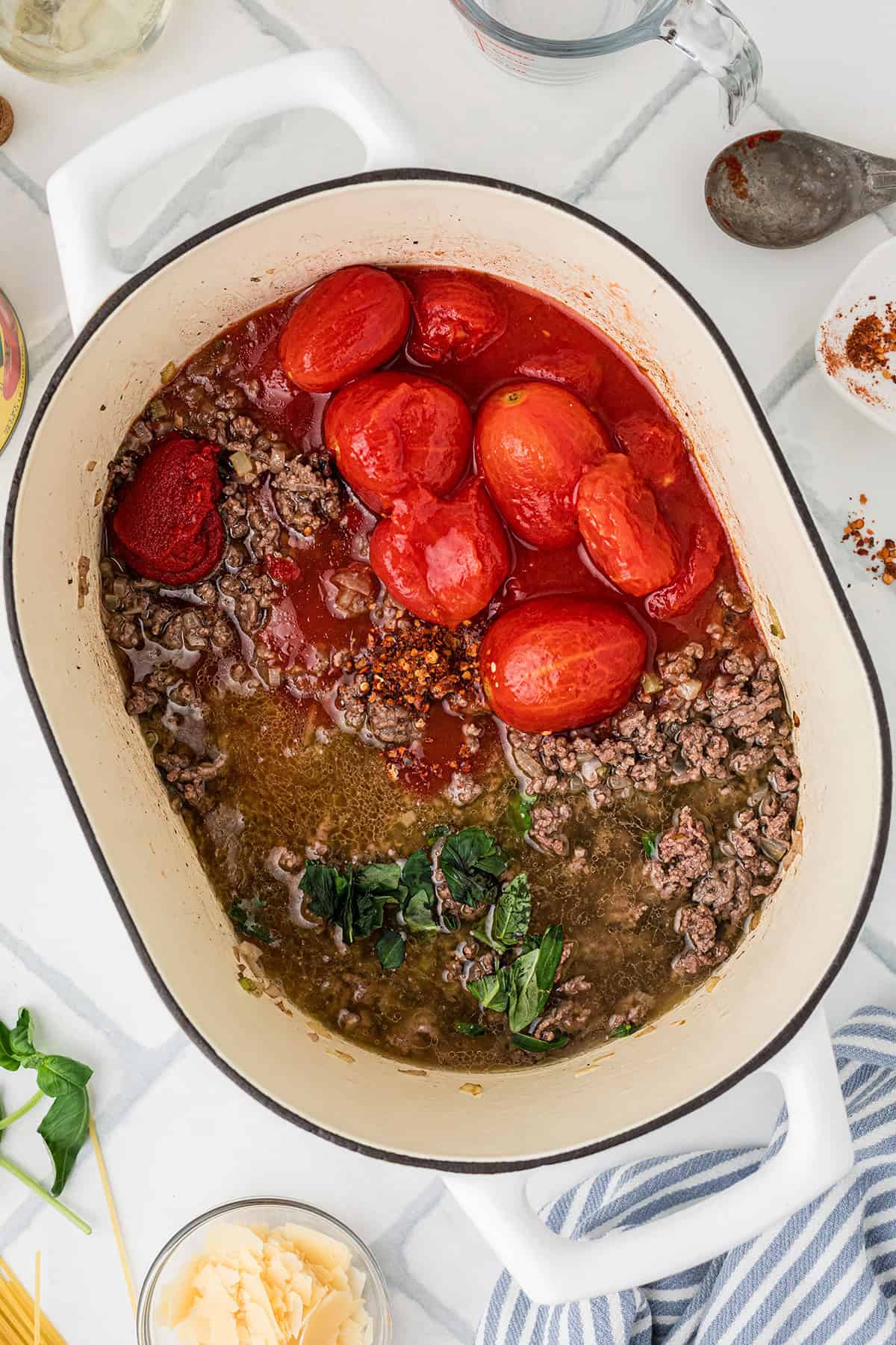 A white pot contains cooked ground beef, whole tomatoes, tomato paste, tomato sauce, herbs, and spices, ready to be mixed for a pasta sauce.