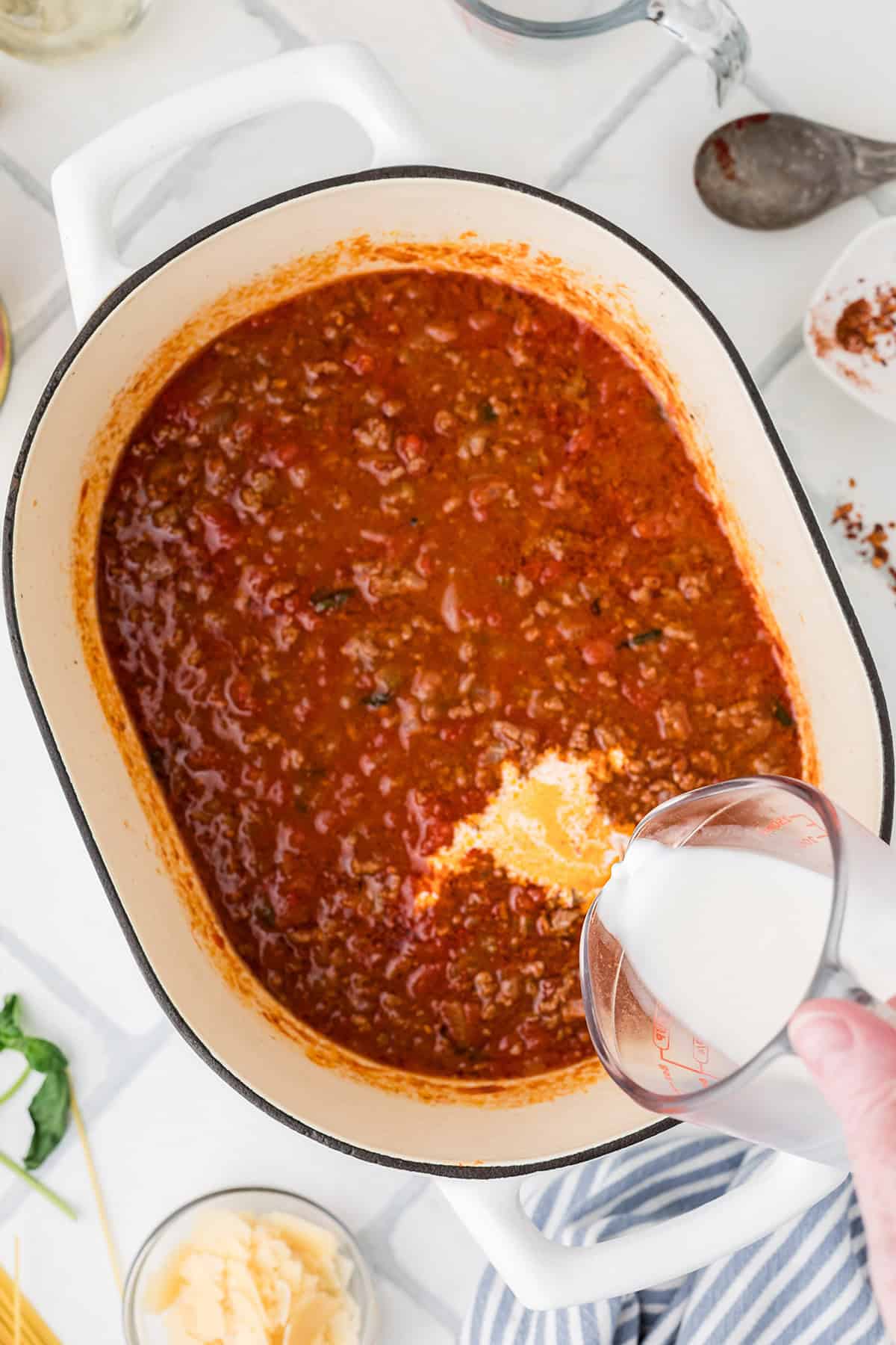 A hand pours half and half from a measuring cup into a pot of red meat sauce on a white stovetop, with cooking utensils and ingredients nearby.