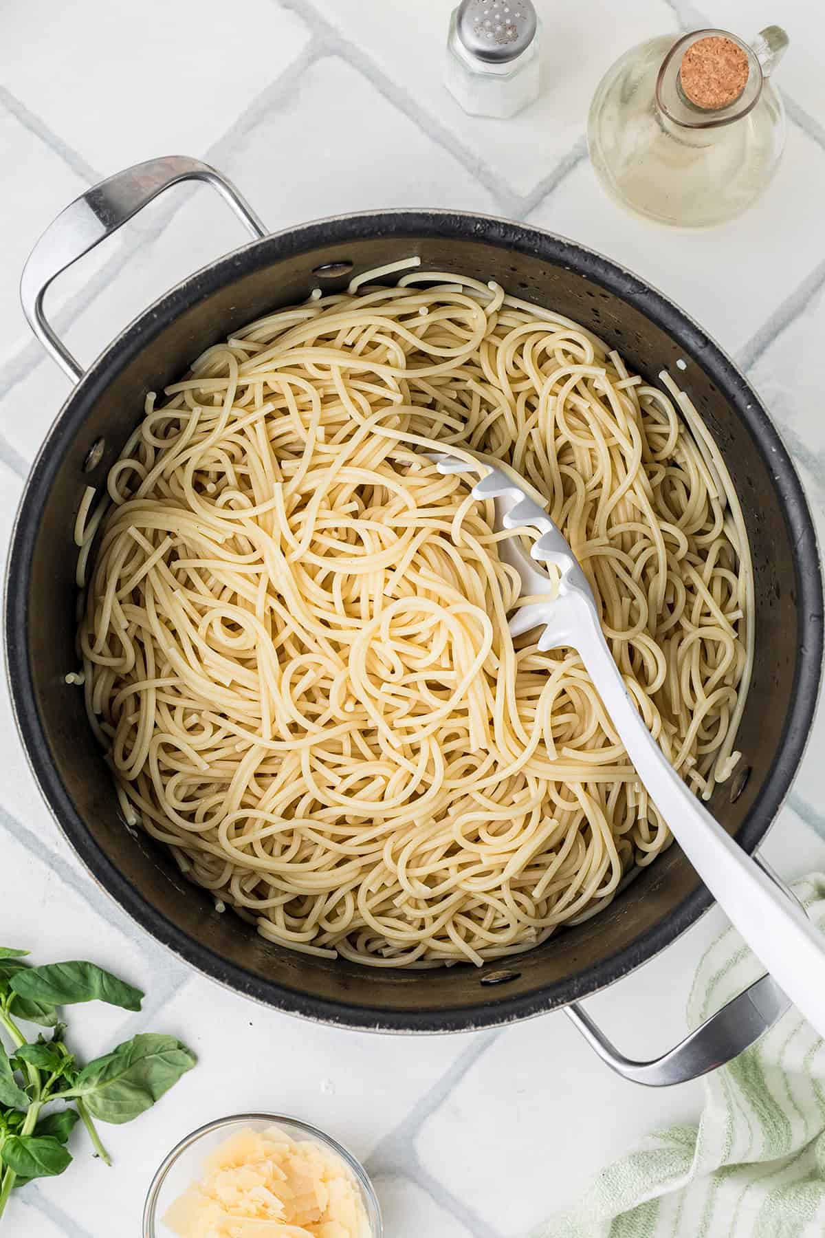 A pot of cooked spaghetti with a pasta serving spoon, surrounded by basil leaves, grated cheese, a salt shaker, and a bottle of oil on a white surface.