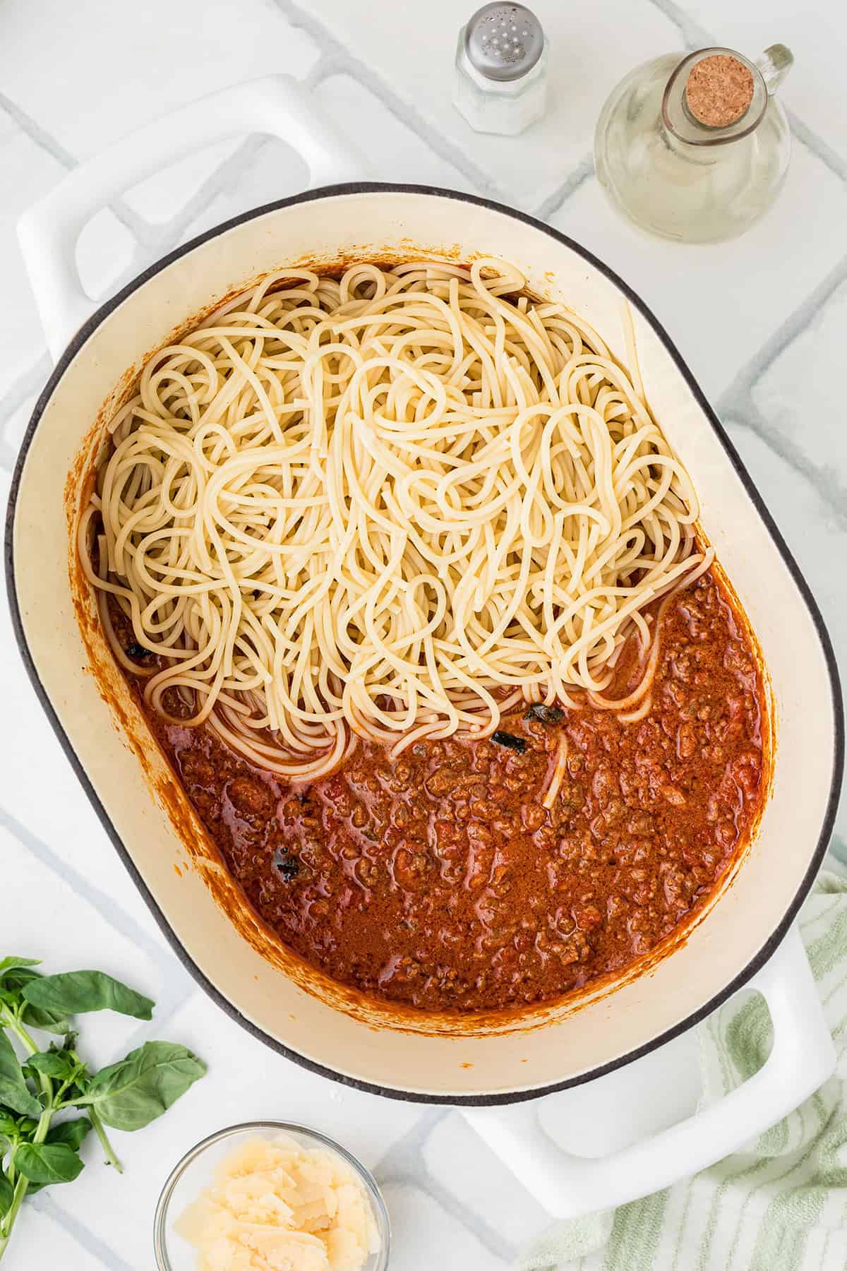 A large white pot containing cooked spaghetti noodles on one side and Bolognese sauce on the other, with various ingredients and seasonings nearby.