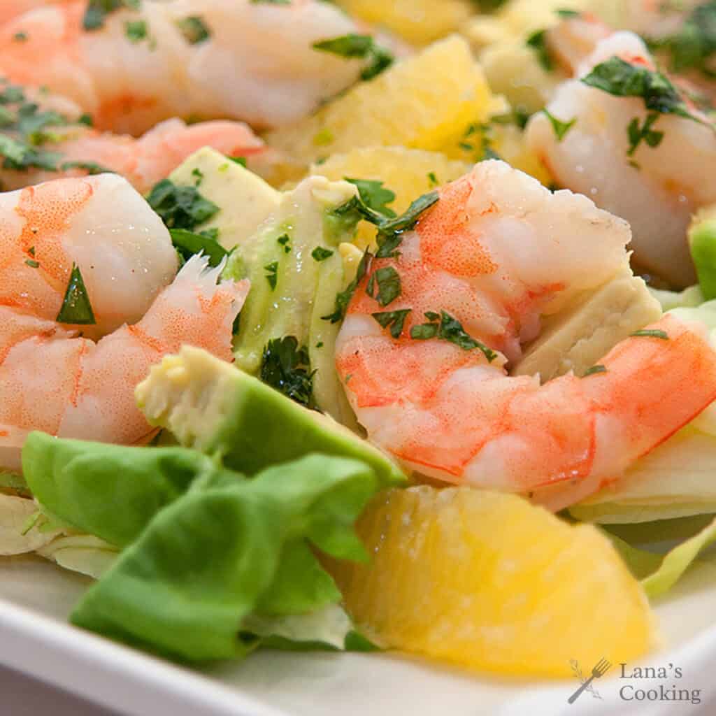 Shrimp salad with avocado, orange slices, fresh greens, and herbs on a white plate.