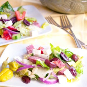 A serving of Greek salad on a white plate with two forks and a bowl in the background.