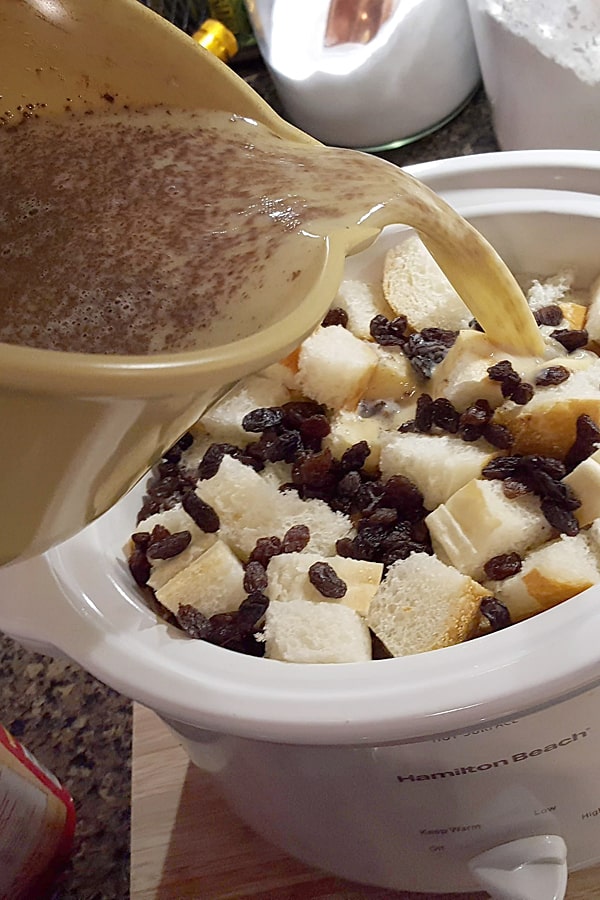 Pouring milk mixture over bread cubes and raisins in the slow cooker.