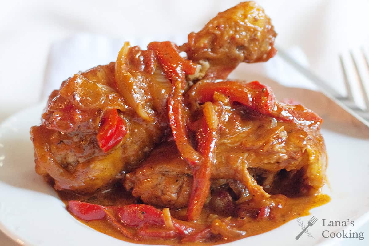 Chicken drumsticks in a rich, red sauce with sliced onions and red peppers on a white plate.