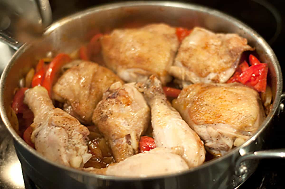 Chicken drumsticks and thighs cooking in a pot with red bell peppers and onions.