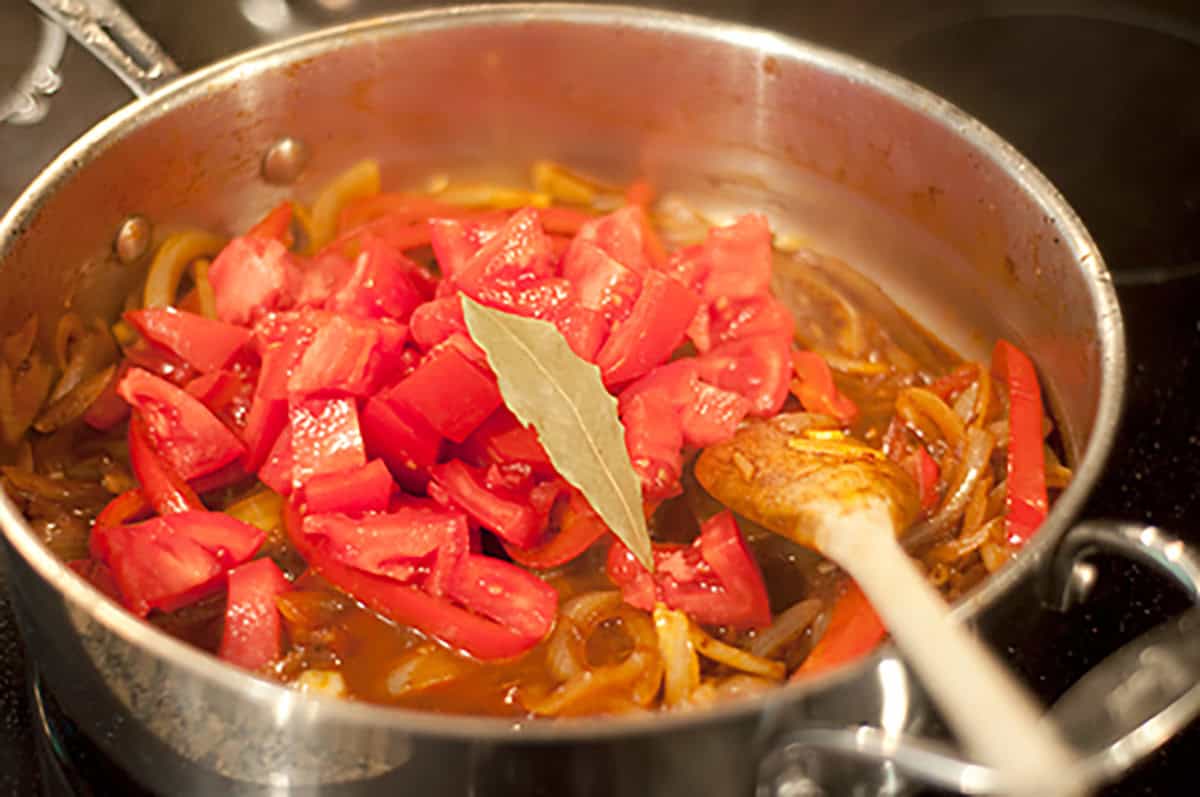 Chopped tomatoes, onions, and a bay leaf cooking in a pot with a wooden spoon.