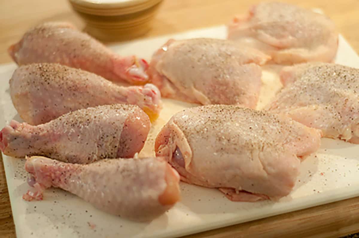 Raw chicken drumsticks and thighs seasoned with pepper on a white cutting board, with a bowl in the background.