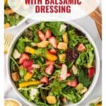 A bowl of strawberry salad with greens, croutons, cheese, and balsamic dressing, viewed from above.