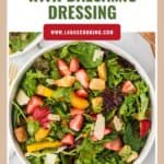 Bowl of strawberry salad with mixed greens, croutons, shaved cheese, and balsamic dressing on a table.