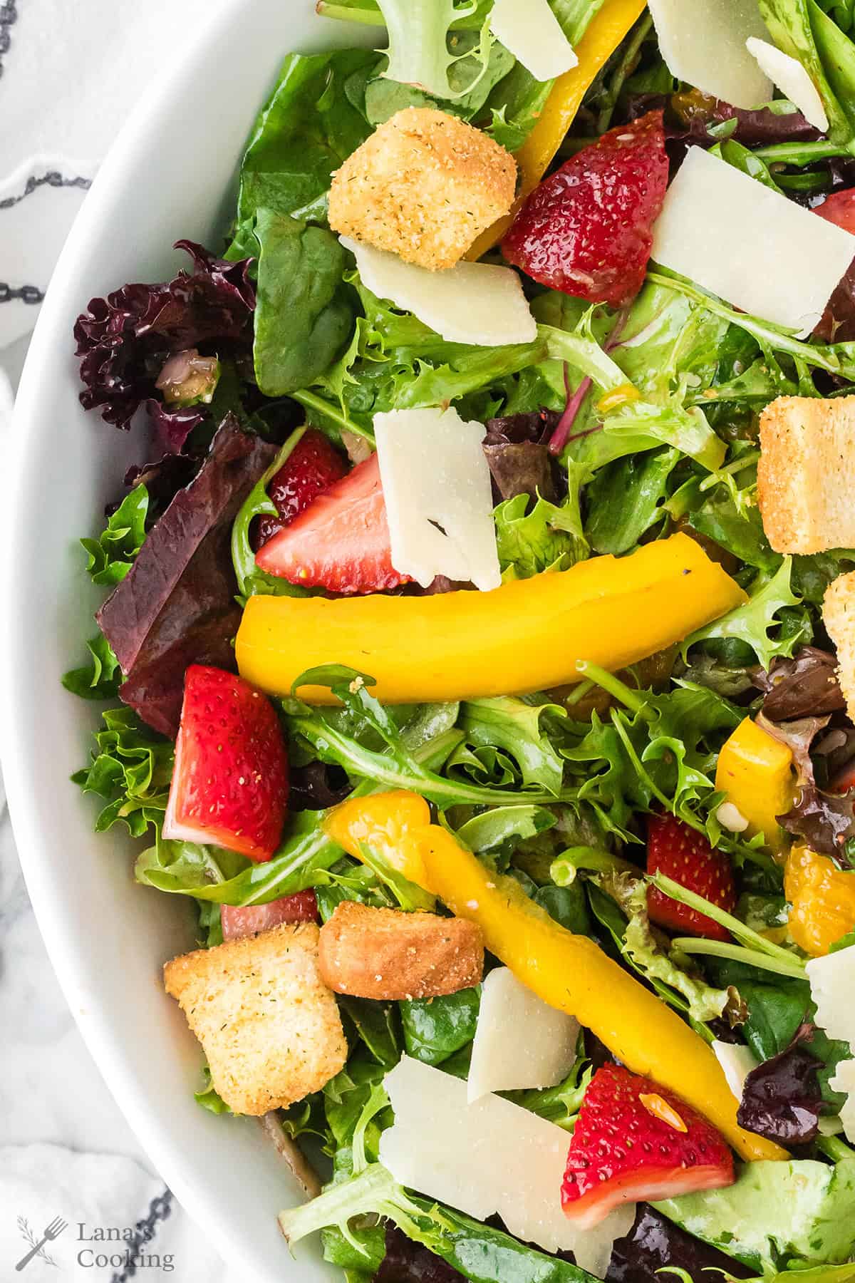 Mixed green salad with sliced strawberries, yellow bell pepper, croutons, and shaved cheese in a white bowl.
