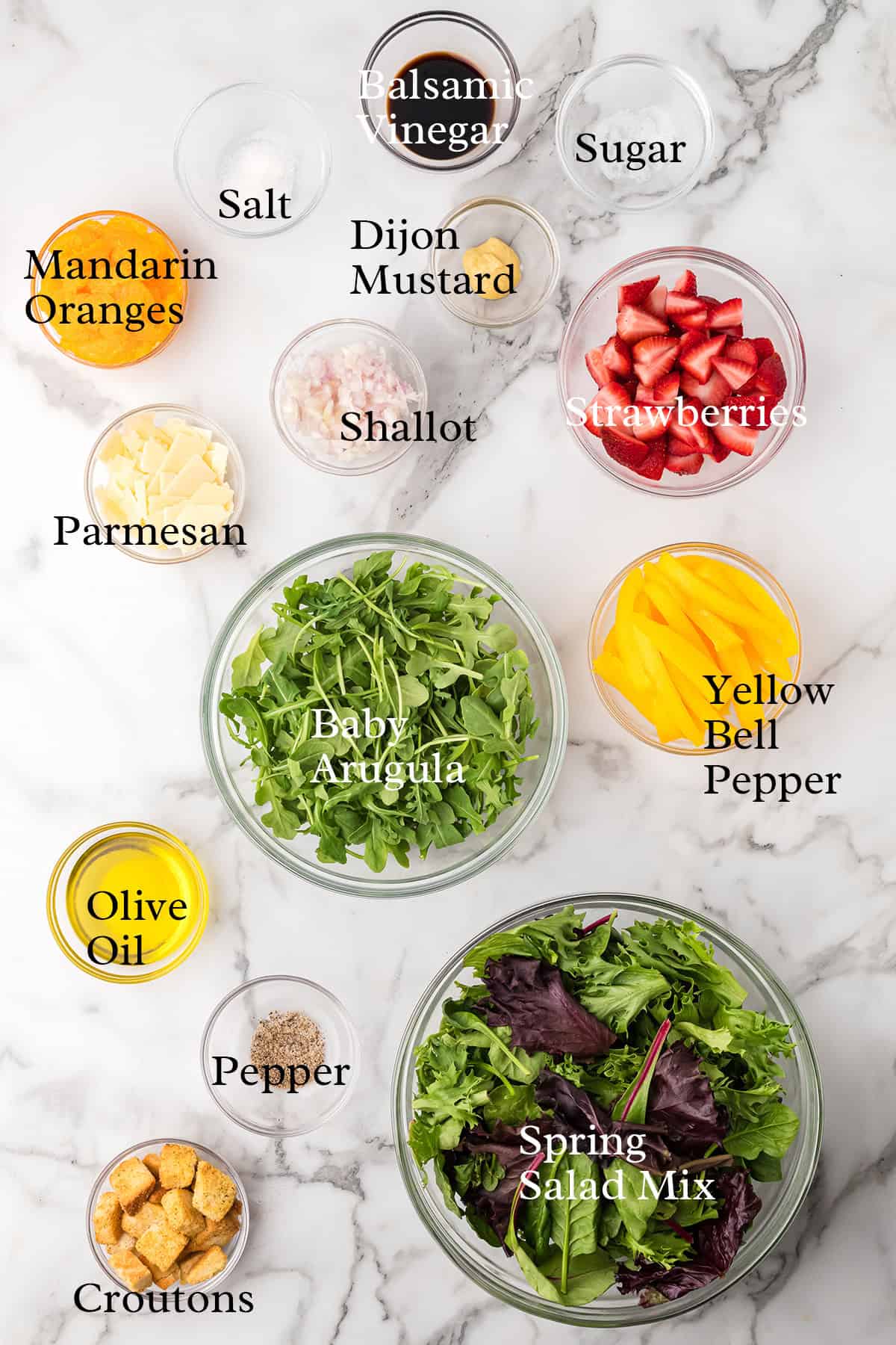 Bowls of salad greens, strawberries, mandarins, bell pepper, parmesan, and salad dressing ingredients on marble.