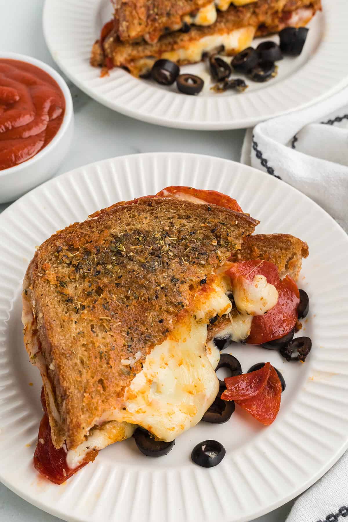 Grilled cheese sandwich with melted cheese, pepperoni, and olives on a white plate, sauce on the side.