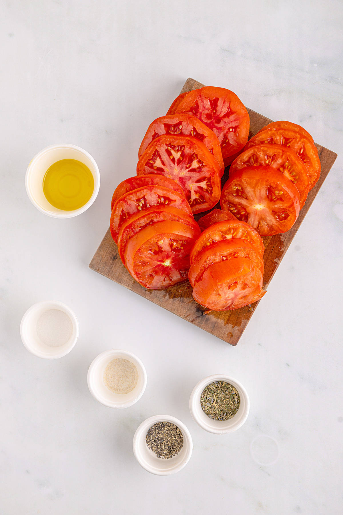 Sliced tomatoes on a wooden board with small bowls of oil, salt, pepper, and herbs on a white surface.