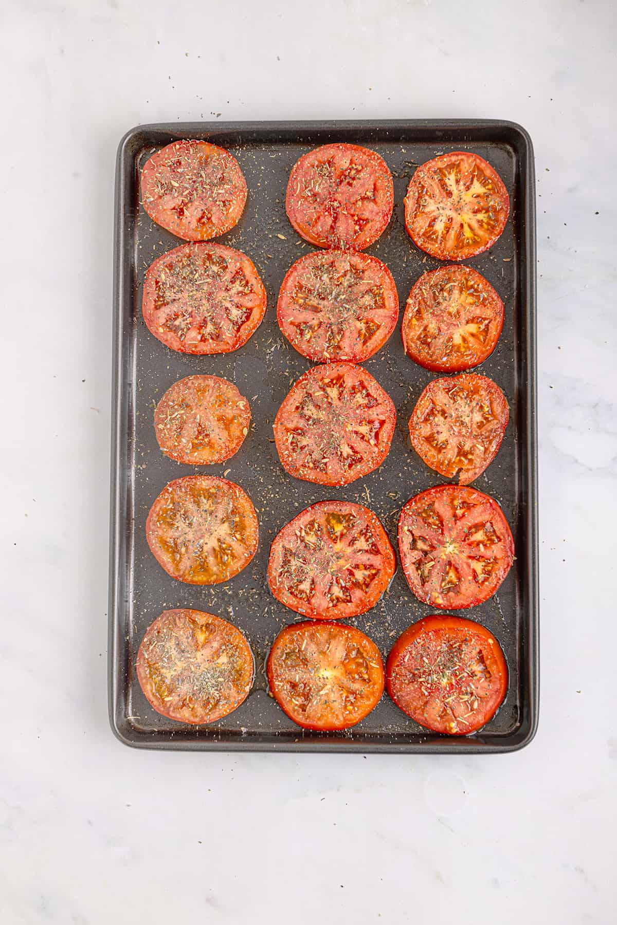 Sliced tomatoes on a baking sheet, sprinkled with herbs and seasonings, ready for roasting.