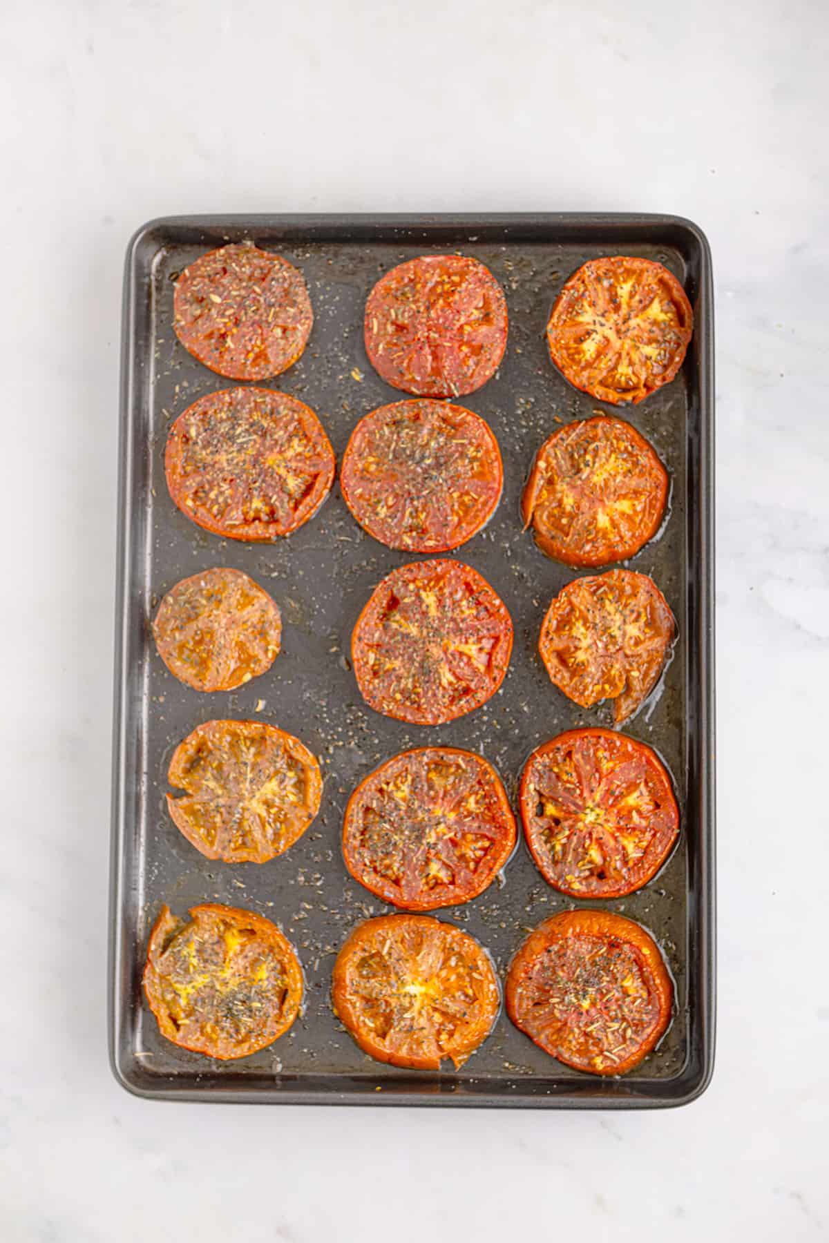 Sliced roasted tomatoes with herbs on a baking sheet, viewed from above on a light surface.