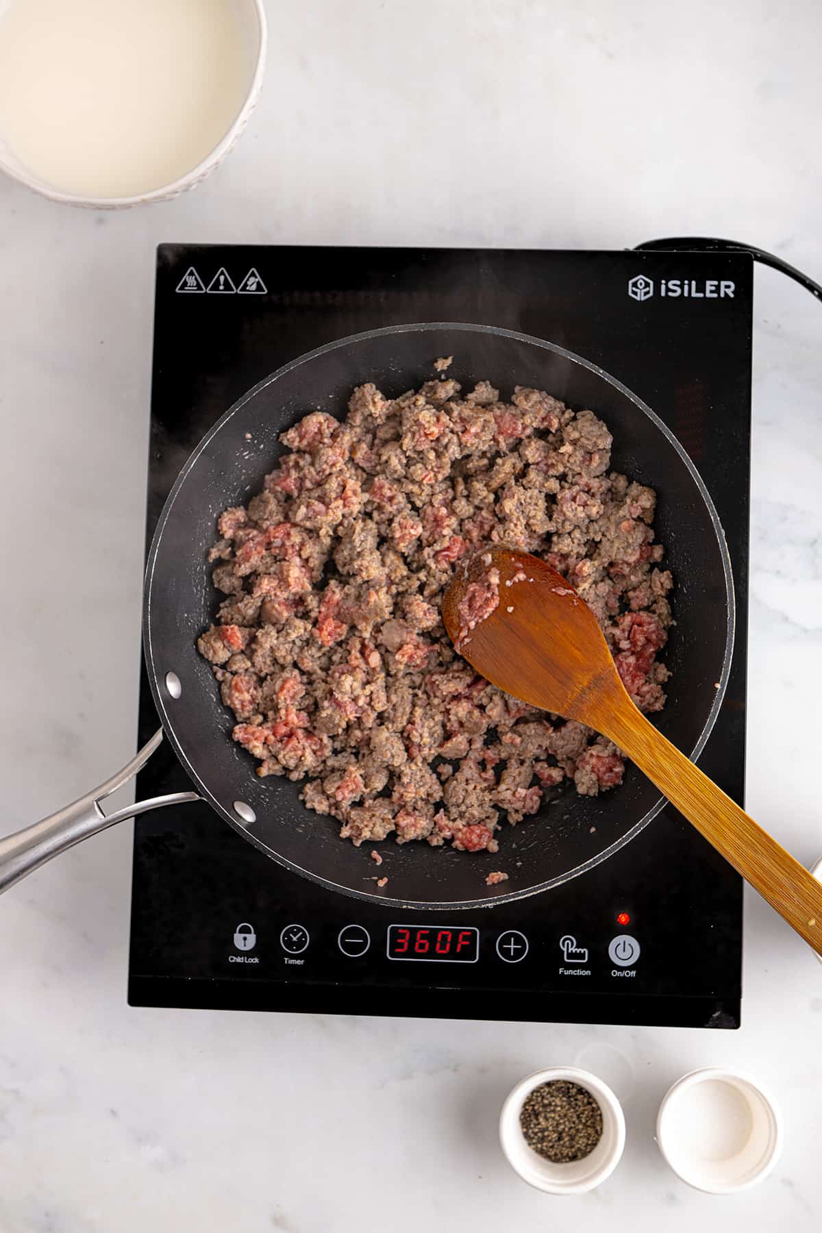 Sausage browning in a skillet on an electric cooktop, with a wooden spoon and seasoning bowls nearby.