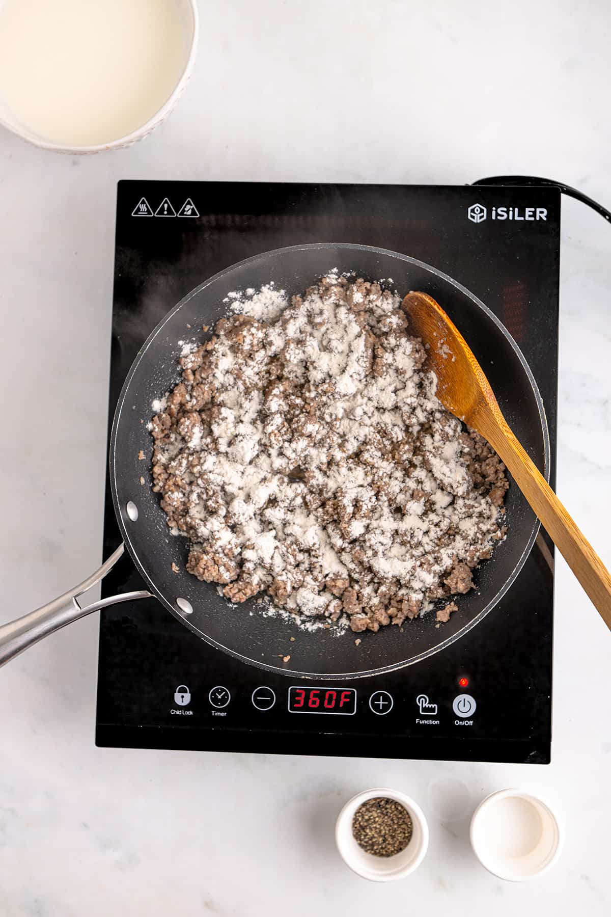 Sausage cooks in a skillet on an induction stove, sprinkled with flour and stirred with a wooden spoon.