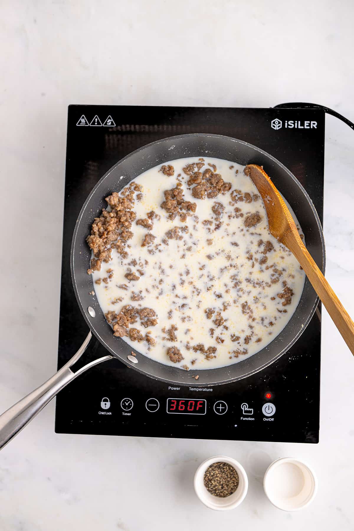 A skillet with sausage and milk is cooking on an induction cooktop, with salt and pepper bowls nearby.