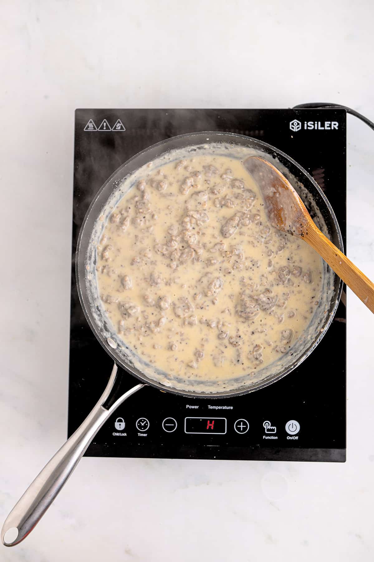 A skillet of creamy sausage gravy cooking on an electric stovetop with a wooden spoon.