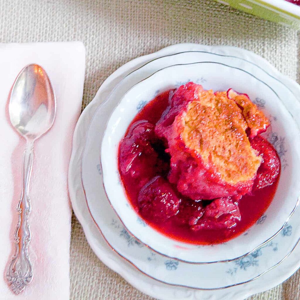 A serving of strawberry cobbler in a china dish with a spoon and napkin alongside.