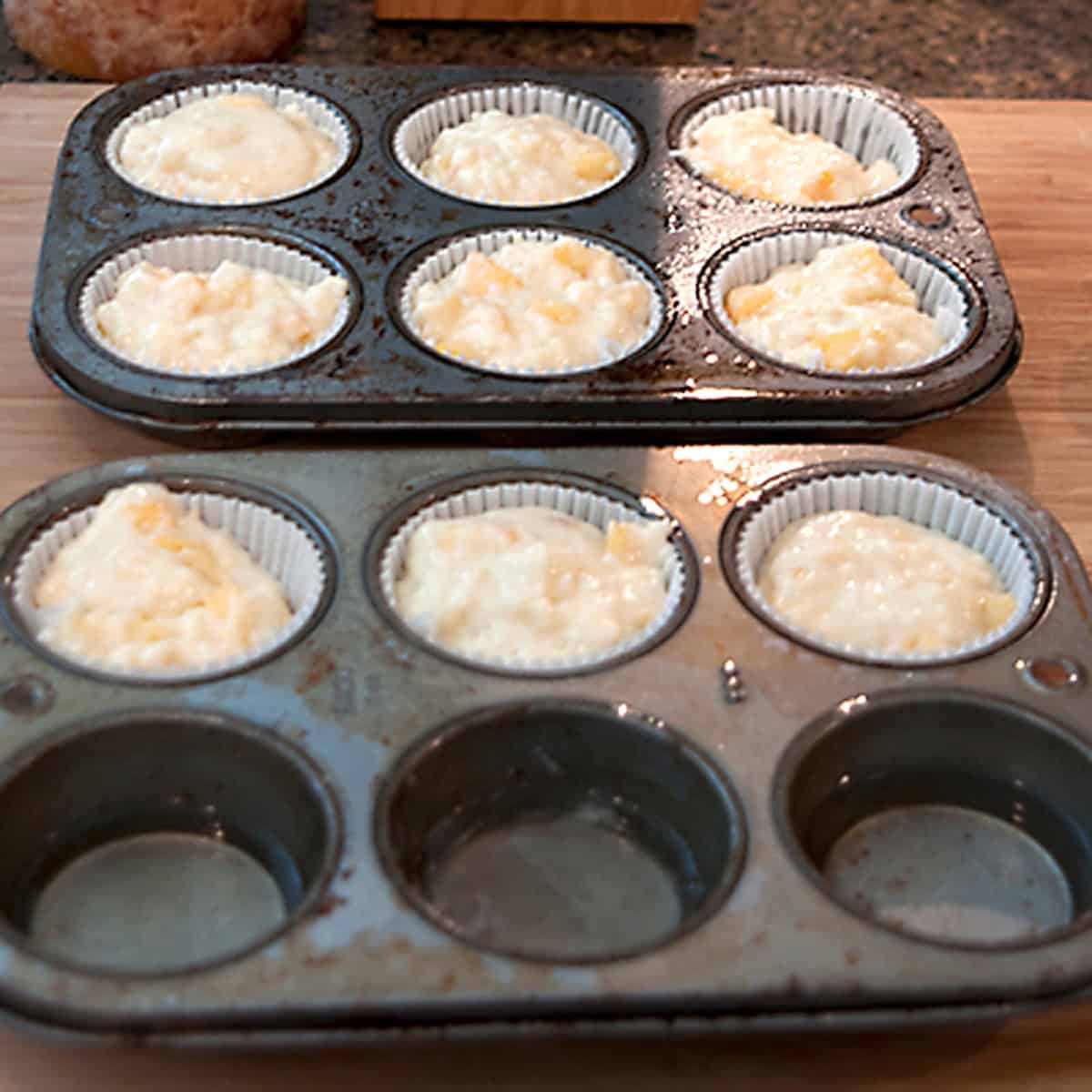 Two muffin trays with paper liners, some filled with batter, on a wooden surface in a kitchen.