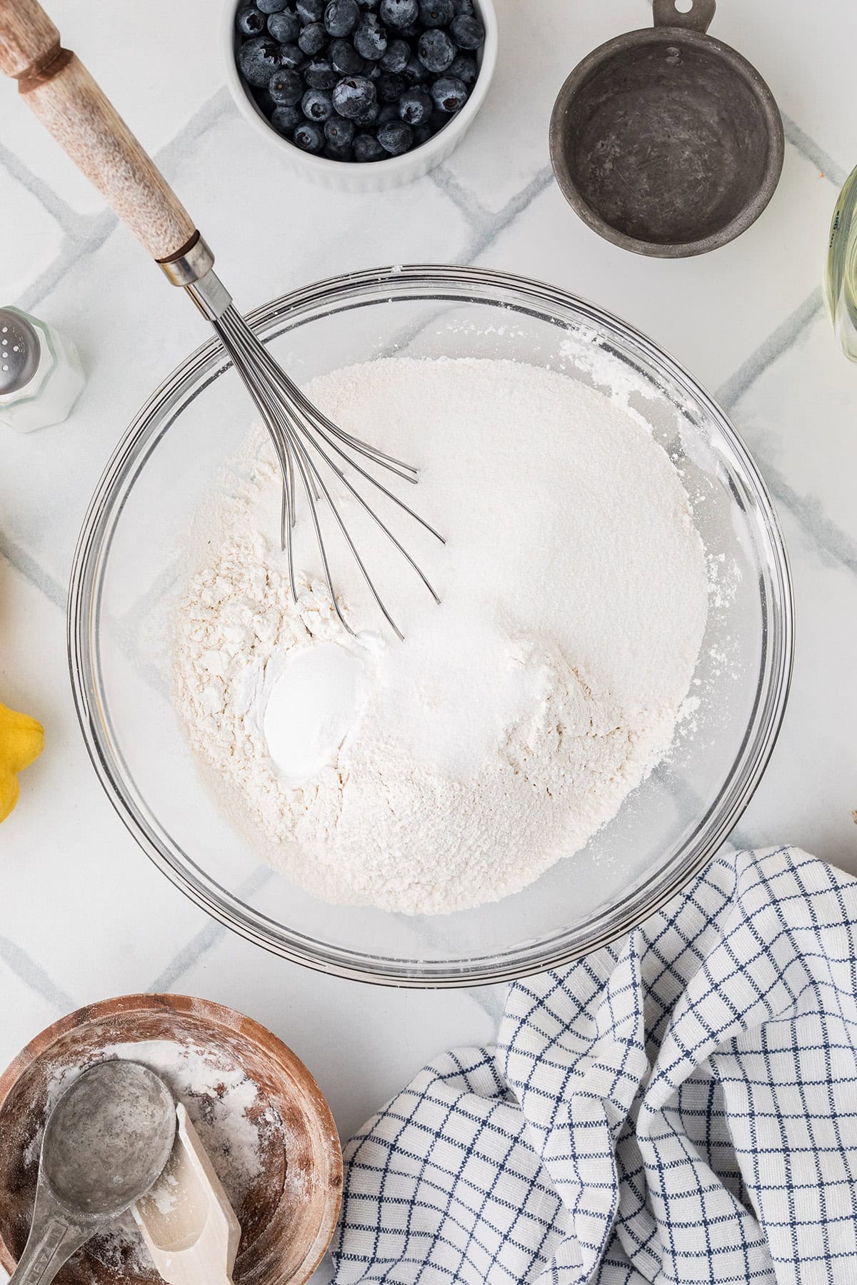 A glass bowl with flour and baking powder being whisked, surrounded by a bowl of blueberries, a measuring cup, a lemon, a checked towel, and other baking tools on a white surface.
