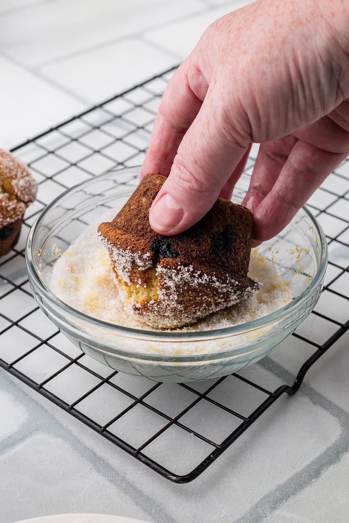 A hand dips a baked muffin into a bowl of sugar on a wire cooling rack.
