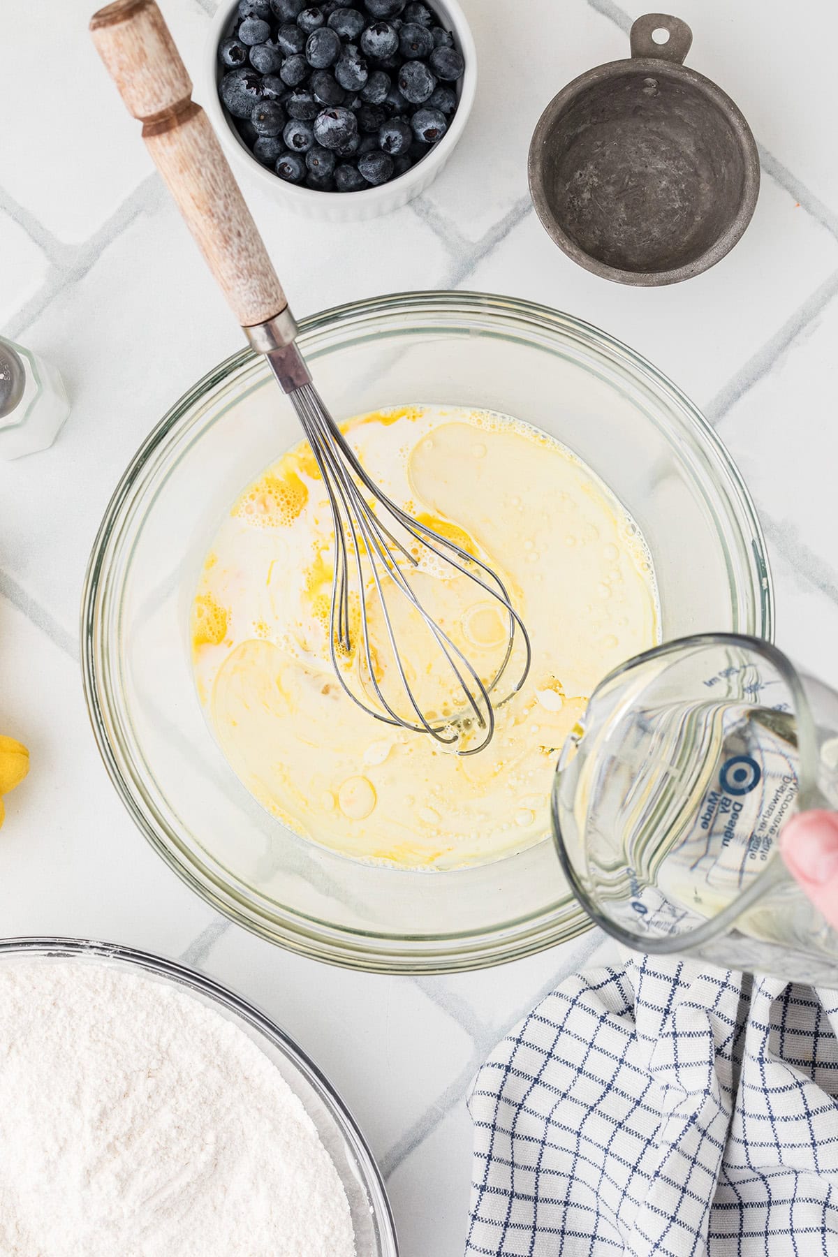 A mixing bowl with eggs and milk being whisked, as oil is poured in. Surrounding the bowl are a towel, flour, blueberries, and a small metal dish.