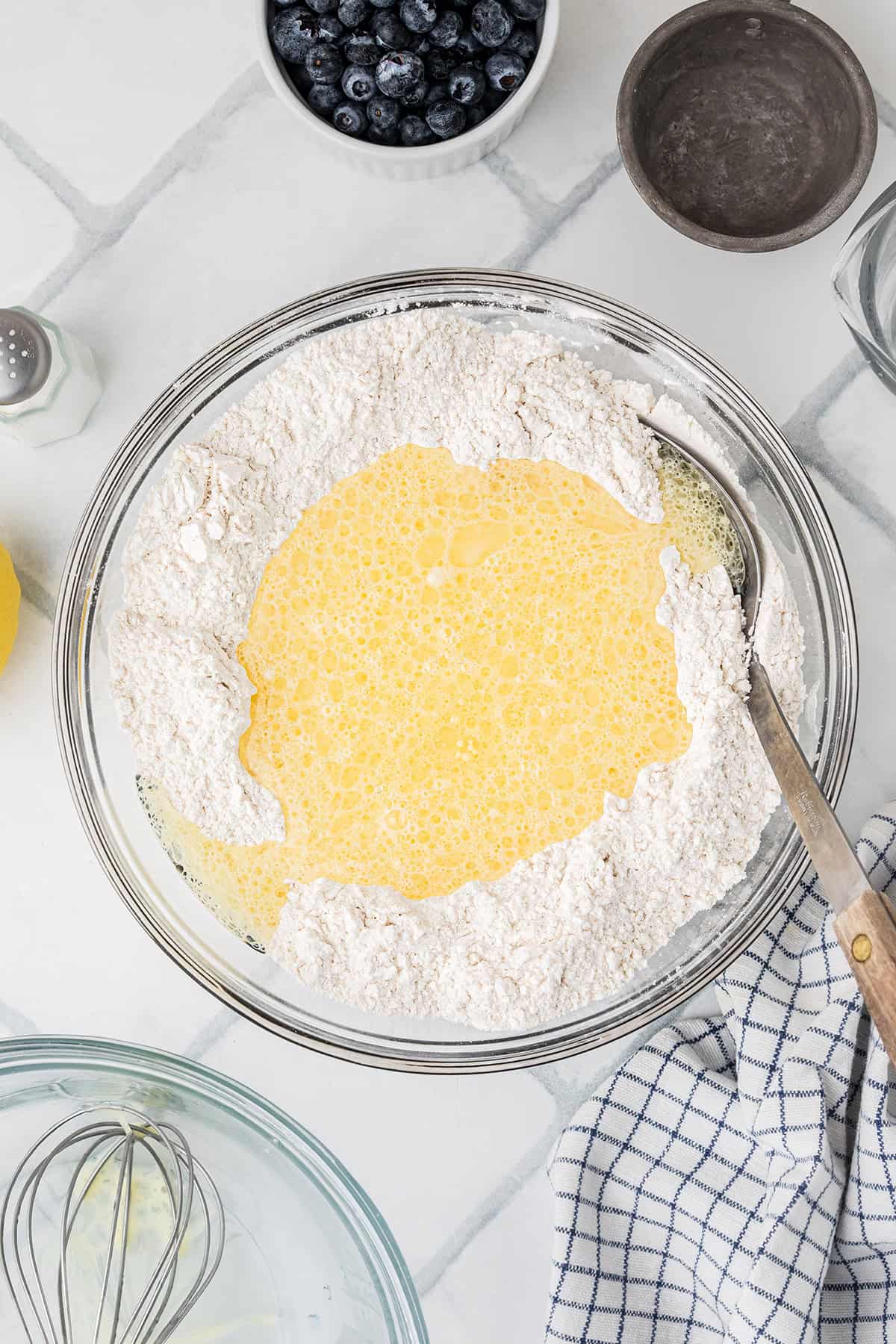 A glass bowl filled with flour and a pool of whisked eggs sits on a tiled counter, surrounded by blueberries, a whisk, and a striped dish towel.