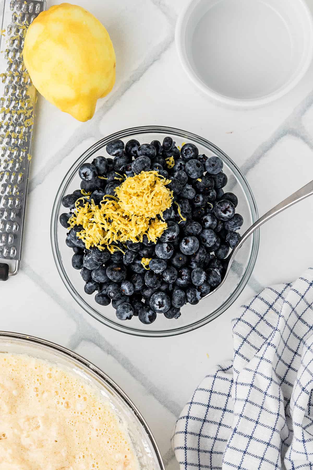 A glass bowl of fresh blueberries topped with lemon zest sits on a countertop beside a whole lemon, a zester, a dish, a linen towel, and a bowl of batter.
