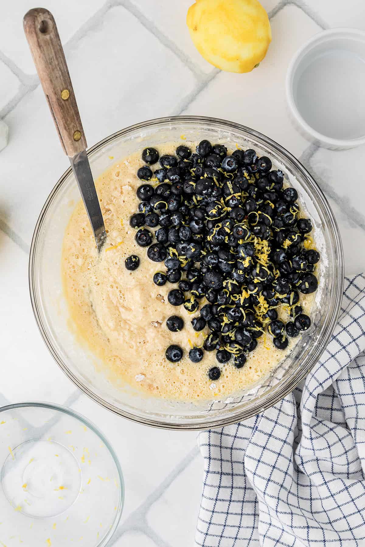 A glass bowl with muffin batter, fresh blueberries, and lemon zest on a tiled surface, with a spoon, lemon, cup, and checkered towel nearby.
