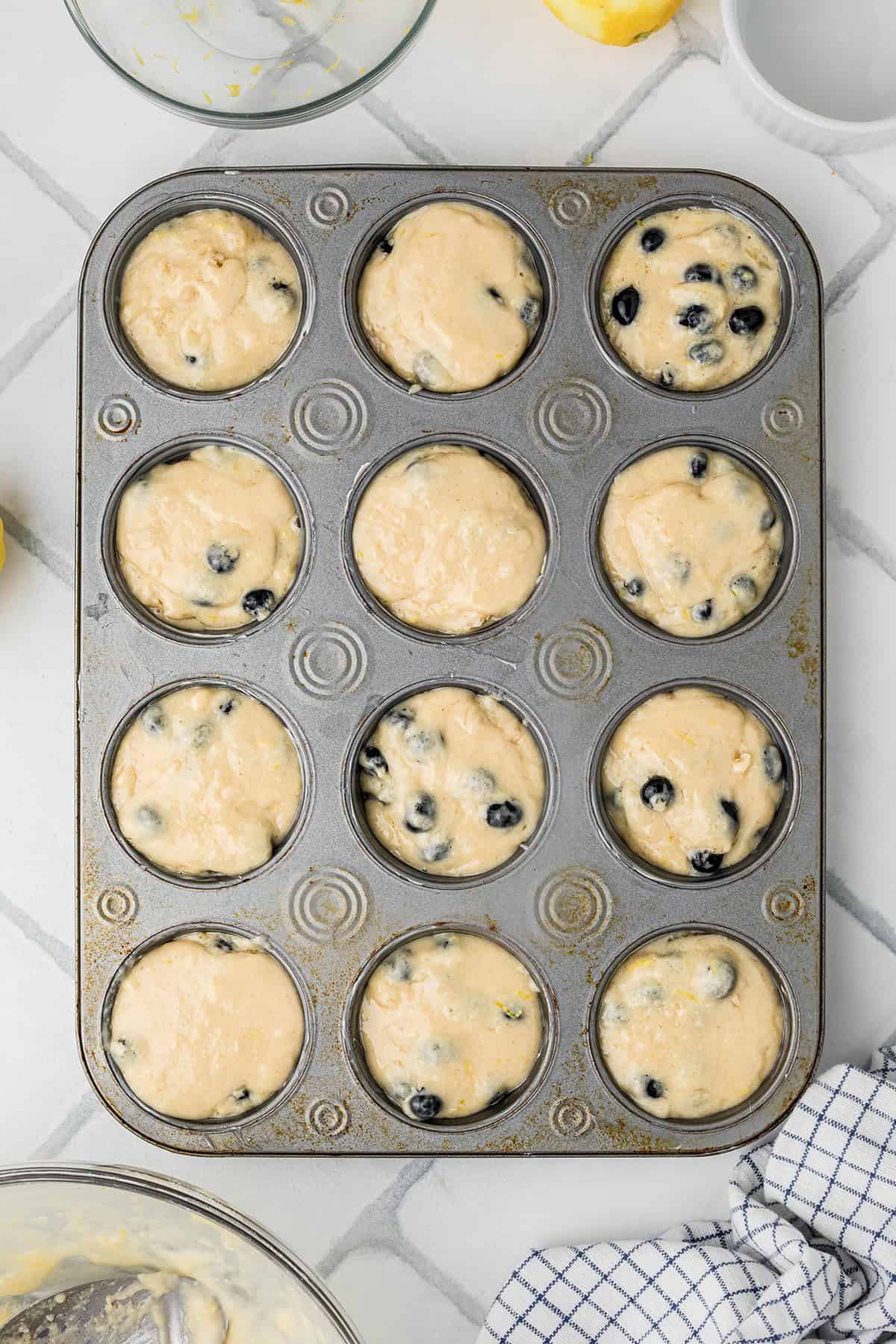 A muffin tin filled with raw muffin batter, some cups containing blueberries, sits on a white tiled countertop.