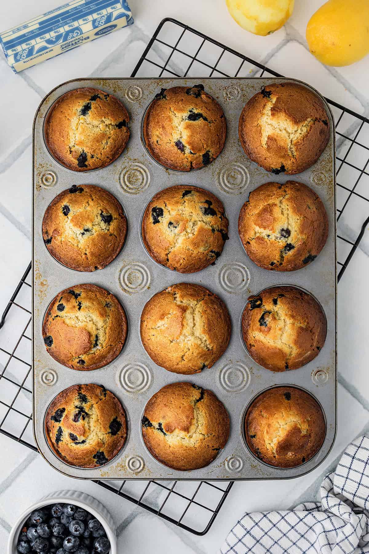 A muffin tin with twelve baked blueberry muffins sits on a cooling rack, surrounded by lemons, butter, fresh blueberries, and a kitchen towel.