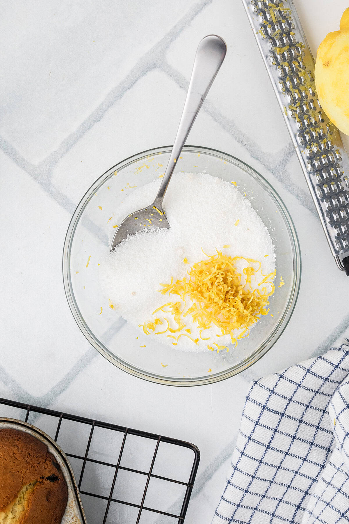 A glass bowl with sugar and lemon zest, a spoon, a grater, a cooling rack with baked goods, and a checkered towel on a white countertop.